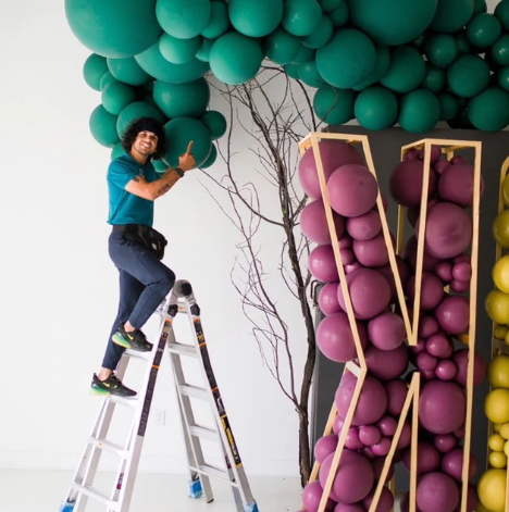 Hired professional Balloon decorator of Bravest Balloons on ladder after finishing a large letter balloon garland and arrangement.